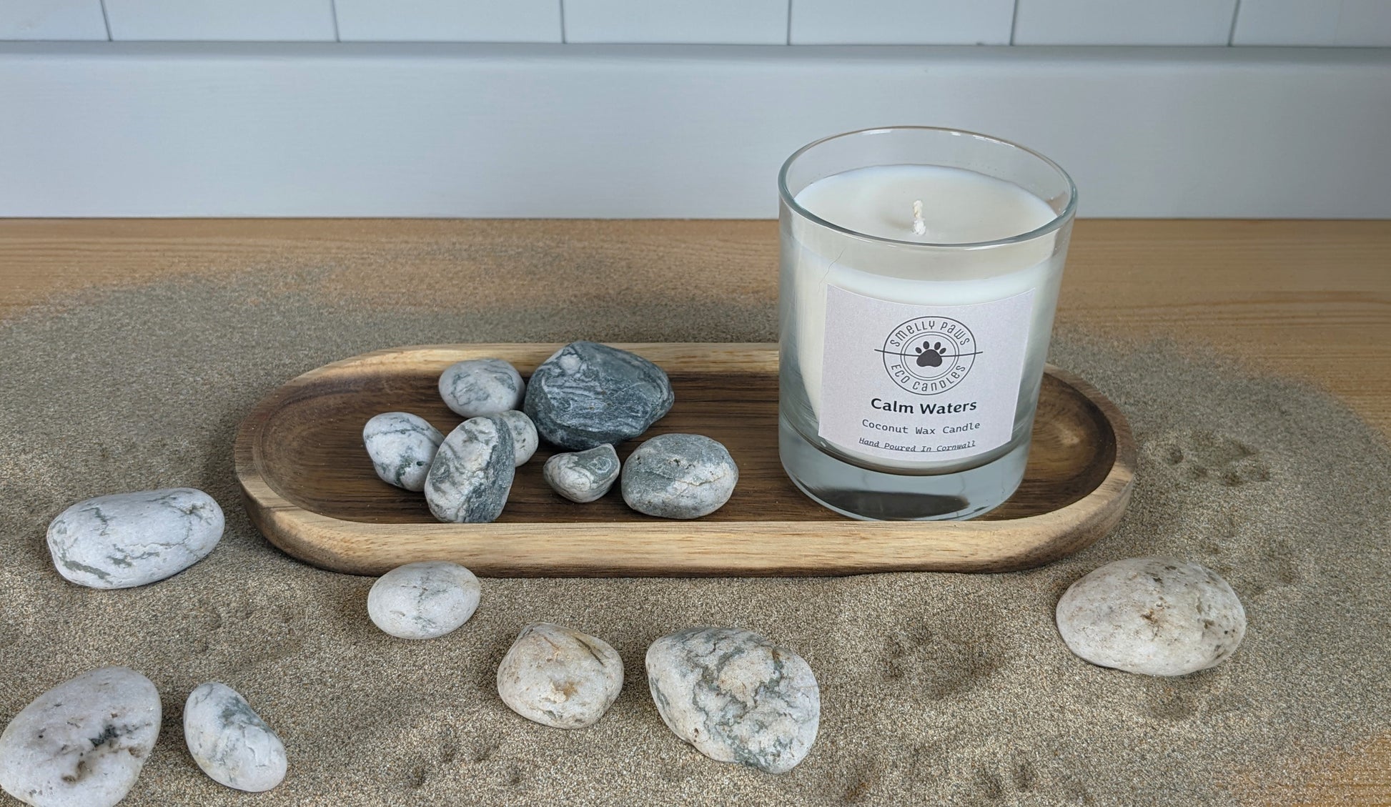 Calm water candle sat on wooden tray surrounded by sand and pebbles 