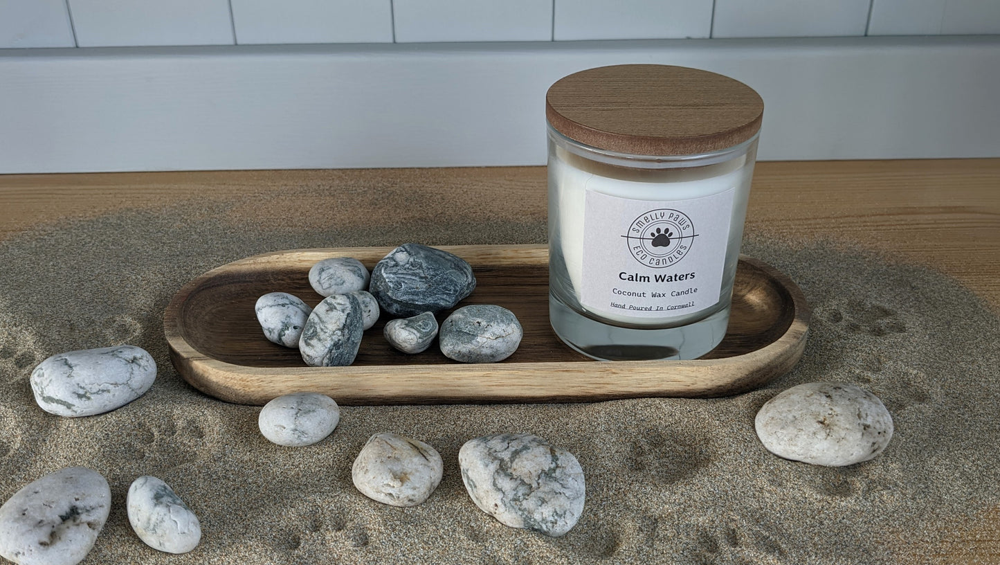 Calm water candle wit wooden lid sat on wooden tray surrounded by sand and pebbles 