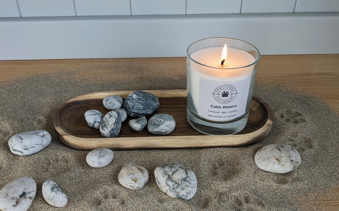 Calm water candle with flame sat on wooden tray surrounded by sand and pebbles 