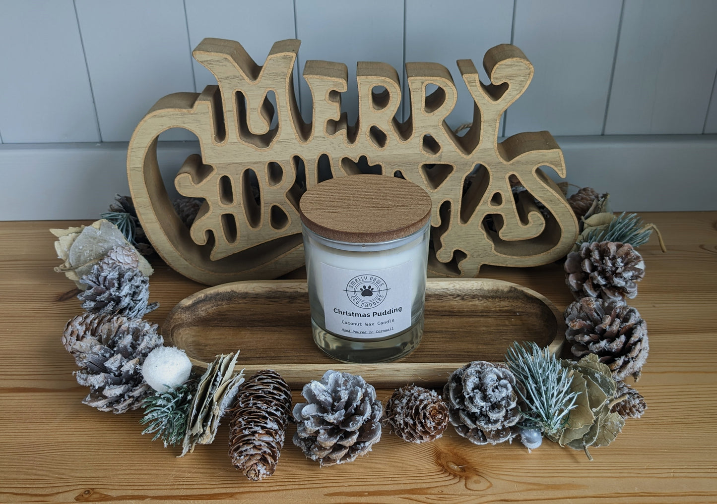 Christmas Pudding candle with wooden lid sat on a wooden tray with pine cones around the front and a merry Christmas sign at the back 