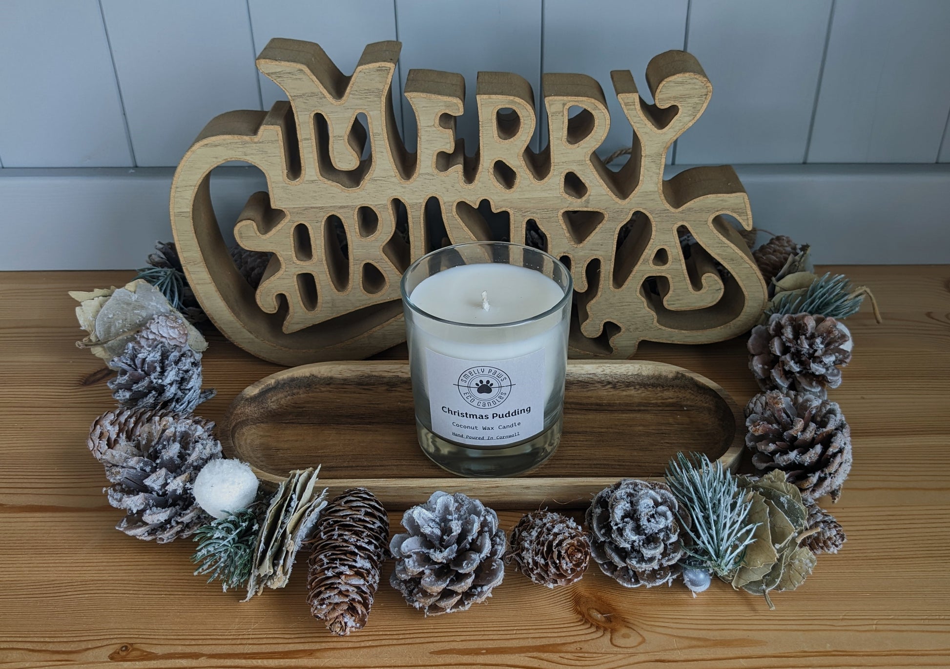 Christmas Pudding candle sat on a wooden tray with pine cones around the front and a merry Christmas sign at the back 