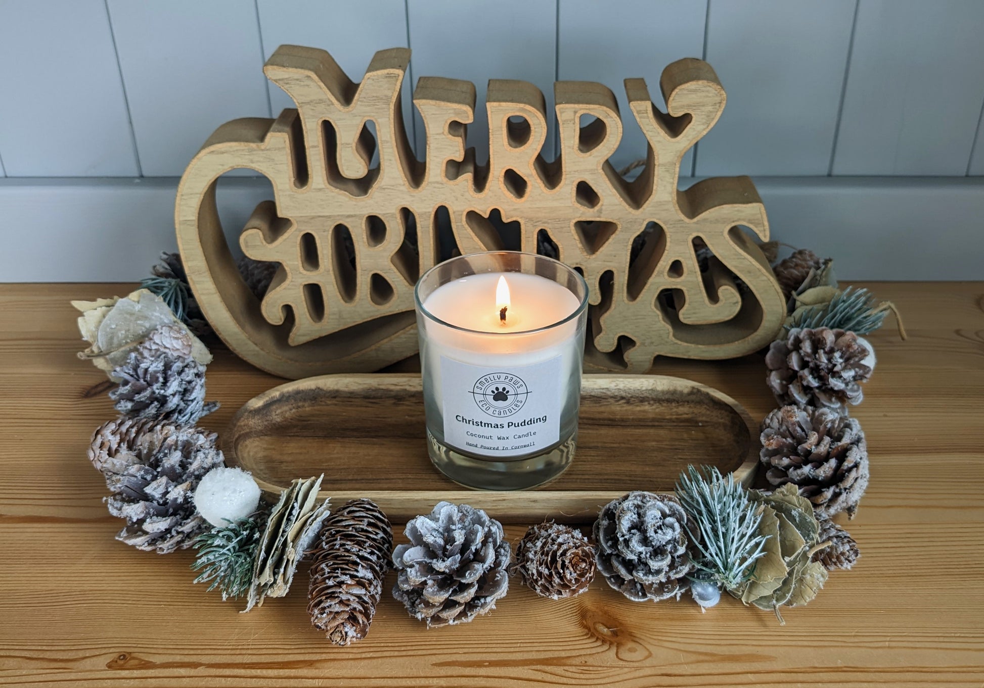 Christmas Pudding candle burning sat on a wooden tray with pine cones around the front and a merry Christmas sign at the back 