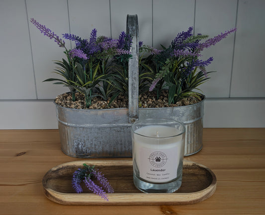 Lavender candle sat on a wooden tray with sprigs of lavender with a metal plant container behind with lavender 