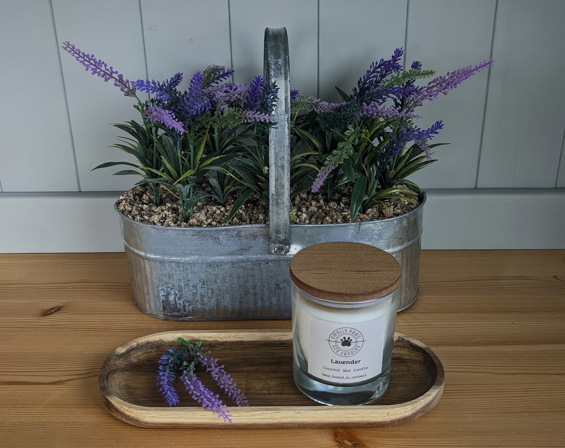 Lavender candle with wooden lid sat on a wooden tray with sprigs of lavender with a metal plant container behind with lavender 