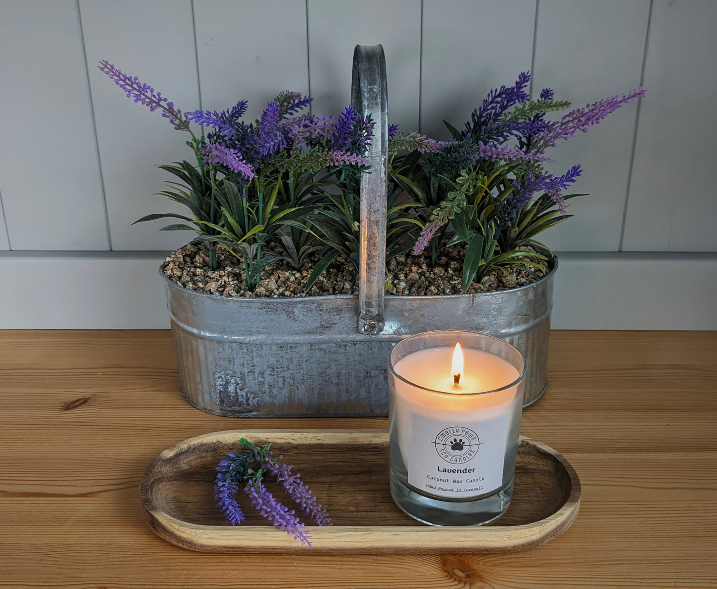 burning Lavender candle sat on a wooden tray with sprigs of lavender with a metal plant container behind with lavender 